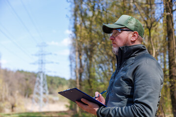 Male engineer monitoring forest health and power line safety during a spring field inspection. Conducting an environmental audit of vegetation near electricity pylons. © A Stock Studio