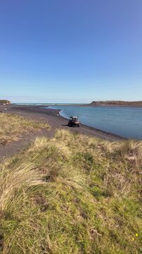 A person drives a utility vehicle near the water's edge on a sandy shore with grassy dunes. Marokopa, Waikato, New Zealand.