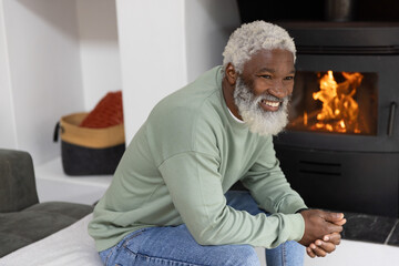 Naklejka premium Senior African American man wearing green sweatshirt sitting in living room by wood stove, smiling