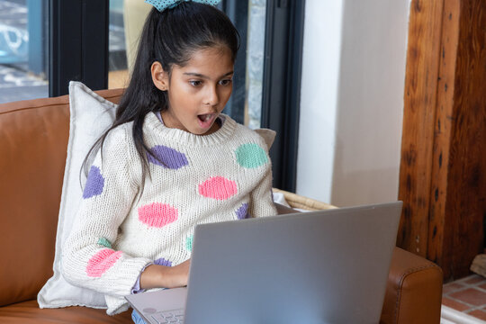 Asian child reacting with surprise on couch using silver laptop at home near dark-glass doors