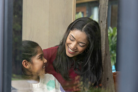 Diverse mother and daughter leaning by large glass window, smiling in knit sweaters