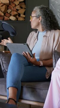 Vertical video: Hearing prompt host with yellow glasses holding tablet on sofa gesturing toward mic