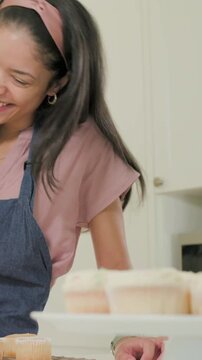 Vertical video: In kitchen girl entering mom guiding pressing piping bag icing cupcakes denim apron