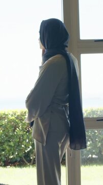 Vertical video: Gazing woman in hijab adjusting scarf, touching chin at window facing sea and hedge