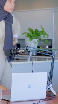 Vertical video: Entering woman in hijab, beige suit tuning mic plugging laptop in kitchen recording