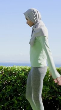 Vertical video: Warming up woman performing stretches on grass by hedge, wearing hijab and sneakers