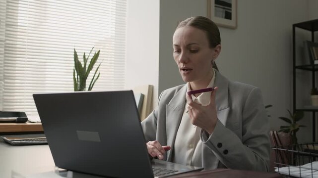 Low angle shot of middle aged businesswoman frowning while talking on phone with client, sitting at table with laptop in office