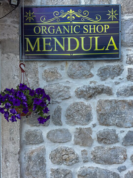  18.04.2026,Perast,Montenegro.Decorative sign reading &ldquo;Organic Shop Mendula&rdquo; mounted on a rustic stone wall, with a hanging pot of purple flowers adding charm.