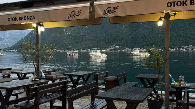  18.04.2026,Perast,Montenegro.Outdoor caf&eacute; seating overlooking calm bay waters with small boats and mountainous scenery in the distance.