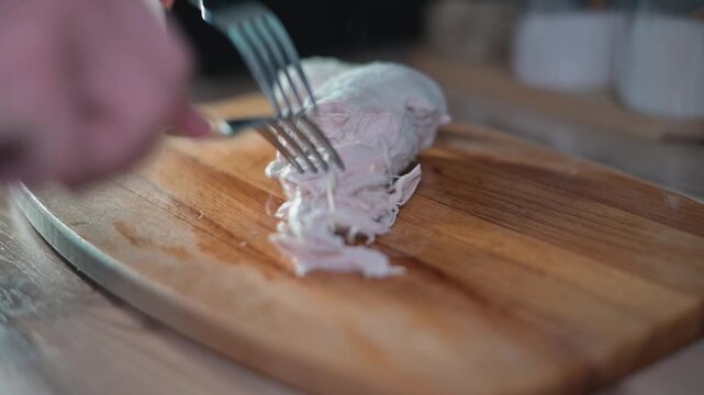 A hand uses two forks to shred cooked chicken on a wooden cutting board. The kitchen setting shows the process of preparing chicken for recipes or meal planning