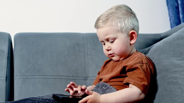 A young child sits on a couch and focuses on a tablet. The room has a simple layout and the light comes from a window. The boy is absorbed in the screen.
