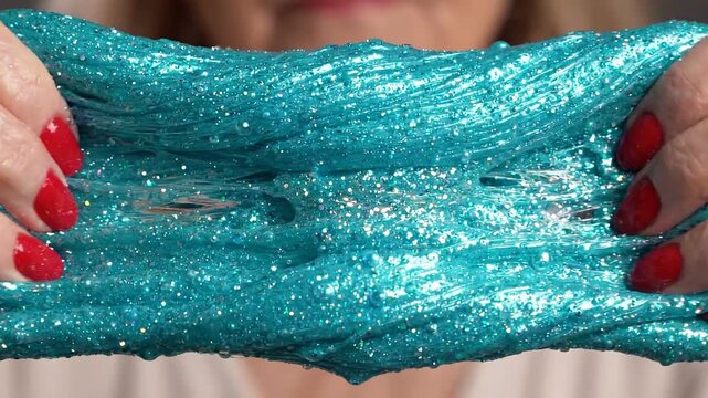 A close-up of hands stretching shimmering blue slime, showcasing its gooey texture, with vibrant red nail polish in focus