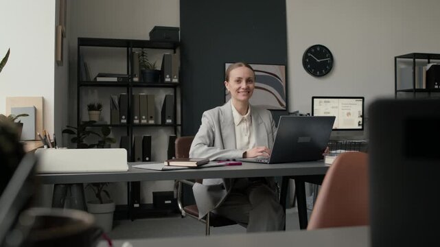Low angle portrait shot of Caucasian businesswoman in suit smiling for camera while sitting at table with laptop in modern office