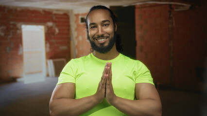 Naklejka premium Man in neon green shirt with hands pressed together in prayer, eyes closed, beard visible, standing in a building under construction; serenity gratitude.
