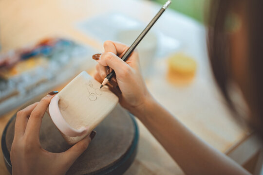 Close-up of female artist&rsquo;s hands sketching design with pencil on handmade ceramic pottery before painting &ndash; creative hobby, traditional craft and artistic expression in calm studio workshop setting
