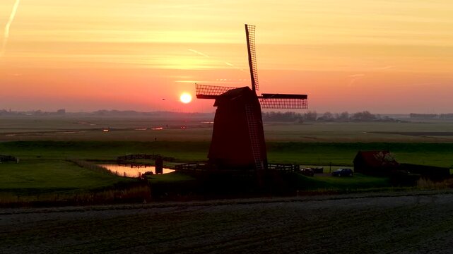 Aerial view of the historic Schermerhorn windmill silhouetted against a vibrant orange sunset over the flat polder landscape and canals in Schermerhorn, North Holland, Netherlands.