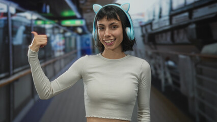 Fototapeta premium Young hispanic woman with cat ear headphones makes finger to lips for silence in a building corridor; quiet.