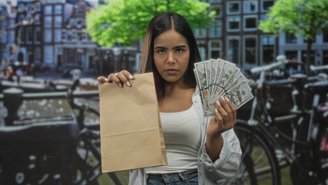 Young woman holds brown paper bag and fans us dollars near bicycles and canal buildings on street; assertive transaction.