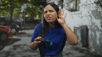 Woman holding gamepad cups her ear with raised hand and puzzled expression on a street next to parked cars and a concrete wall  curiosity challenge. © Krakenimages.com