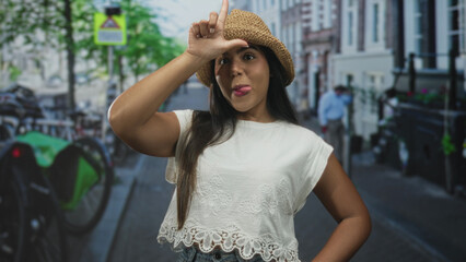 Fototapeta premium Young hispanic woman forms l with hand on forehead while sticking out tongue, wearing straw hat and white lace top on a city street; playful mischief.