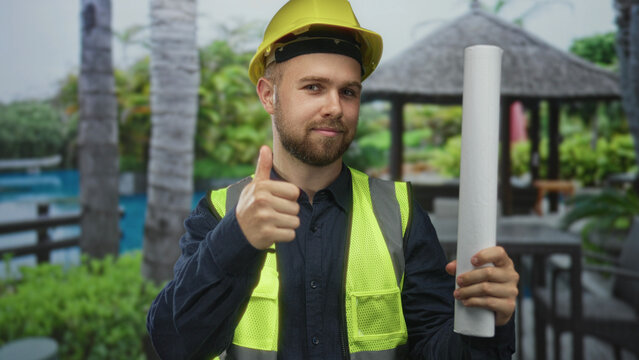 Man construction worker wearing yellow hardhat and high vis vest holding a rolled blueprint and giving thumbs up at building; confidence safety.