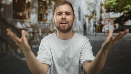 Fototapeta premium Man with open hands gesturing in white tshirt on street with blurred buildings and trees; thoughtful reflection.