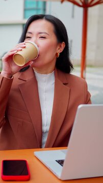 Asian woman manager business entrepreneur working on laptop pc computer at outdoor cafe. Smiling mature businesswoman holding coffee cup, urban modern background, remote work lifestyle. Vertical