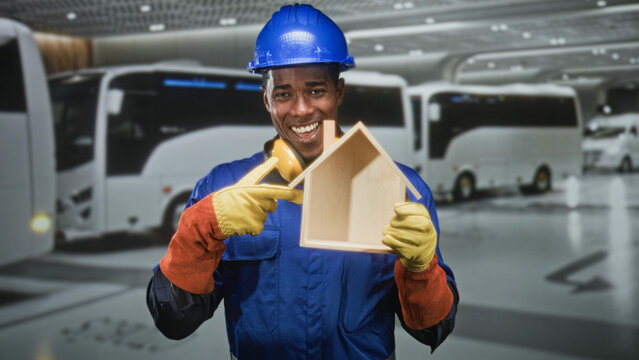 Man builder in hardhat points at wooden house model inside bus terminal building while smiling and wearing yellow gloves and earmuffs; pride duty.