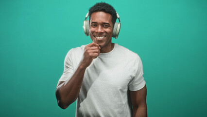 Naklejka premium African american man wearing over ear headphones, smiling with right hand to chin and relaxed posture against teal seamless studio backdrop; joyful confidence.