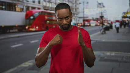 Fototapeta premium Man clasping hands in a red t shirt in front of a red double decker bus on a busy city street with blurred pedestrians and vehicles; determination.