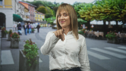 Fototapeta premium Woman pointing finger in white shirt on street near crosswalk and outdoor cafe tables; warm confidence.