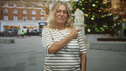 Fototapeta premium Senior woman points finger at decorated christmas tree on street, visible hand gesture, wearing striped shirt and glasses, outdoor plaza scene; skepticism.
