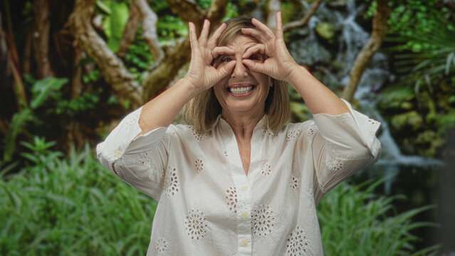 Caucasian middle age woman in a white blouse frames her eyes with hand binoculars in a lush forest setting; exploration joy.