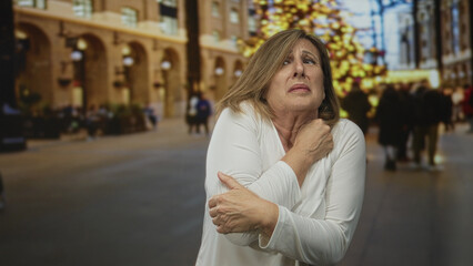 Fototapeta premium Woman clutch chest with right hand and hug left arm on crowded city street under festive christmas lights; pain distress anxiety.
