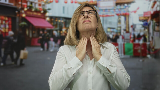 Woman clutches throat in busy urban street scene by storefront wearing white shirt amidst blurred crowd; panic and urgency.