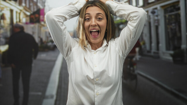 Woman holding hands on head and grimacing in a busy city street while pedestrians pass and storefronts line the sidewalk; stress frustration.
