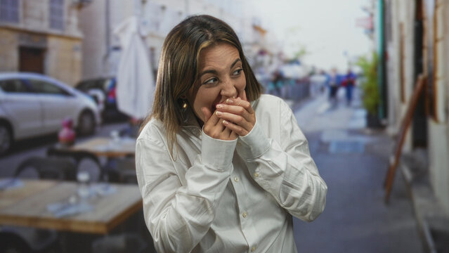 Woman with hands covering mouth and fingers clasped, eyes closed, wearing white shirt on a restaurant terrace outdoors by sidewalk tables and umbrellas; surprise reaction.