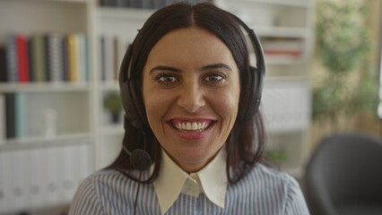 Woman wearing headset and microphone speaking and smiling, visible teeth, striped blouse, seated at desk inside office building with bookshelves and plant  warmth connection. © Krakenimages.com