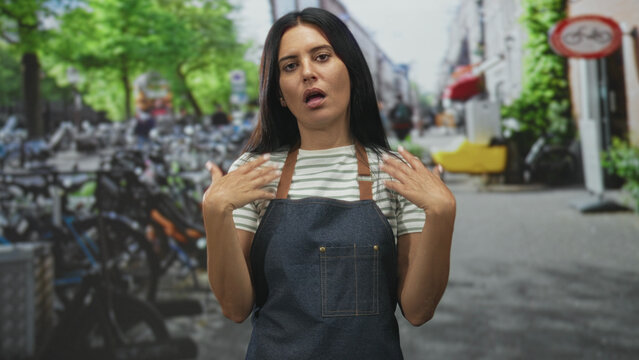 Woman in denim apron flips hair with right hand, hands visible, gesturing dismissively by parked bicycles on street; exasperation dismissal.