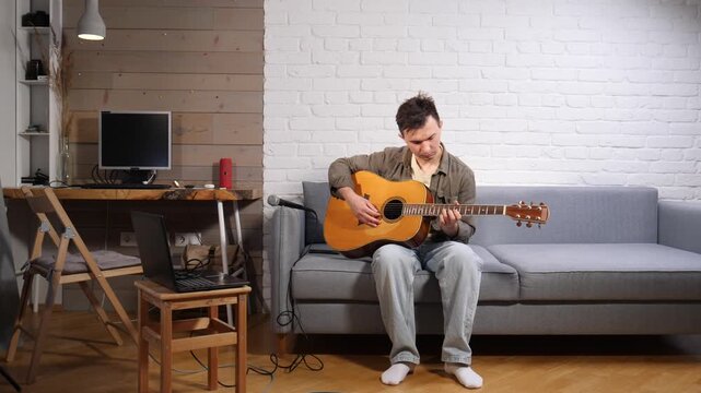Young man relaxing on a sofa while strumming an acoustic guitar, engaging in a creative hobby and practicing music at home with a laptop nearby for learning and recording