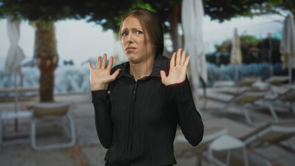 Fototapeta premium Woman with palms up hands raised and tense fingers near chest in studio poolside set with lounge chairs and umbrellas; unease.