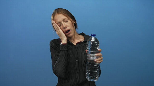 Woman holding a large water bottle with hand on cheek yawning in blue studio; tiredness dehydration thirst.