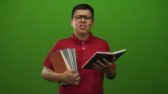 Young hispanic man holding textbooks and squinting while reading a notebook in a studio with green backdrop; study frustration.