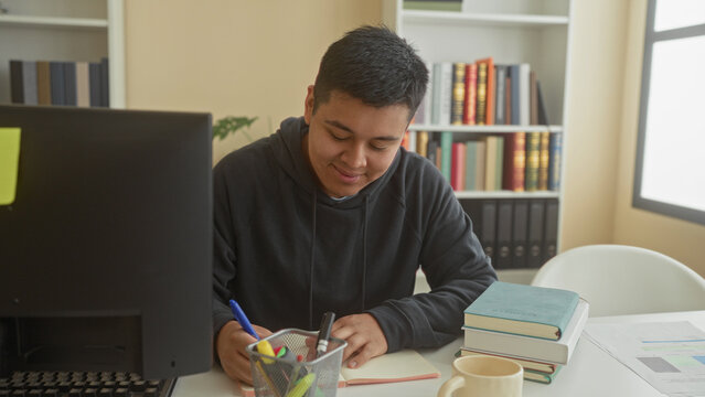 Man writing with pen at desk in library, stack of books, monitor and papers visible, smiling while studying and taking notes; focused study.