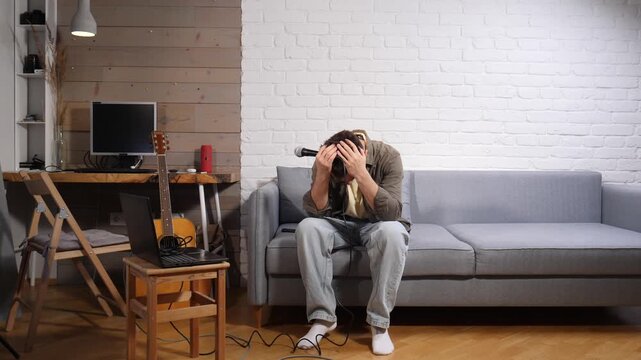 Young man sitting on a gray couch with his face buried in his hands and a microphone nearby, feeling overwhelmed and exhausted from creative block and emotional distress at home