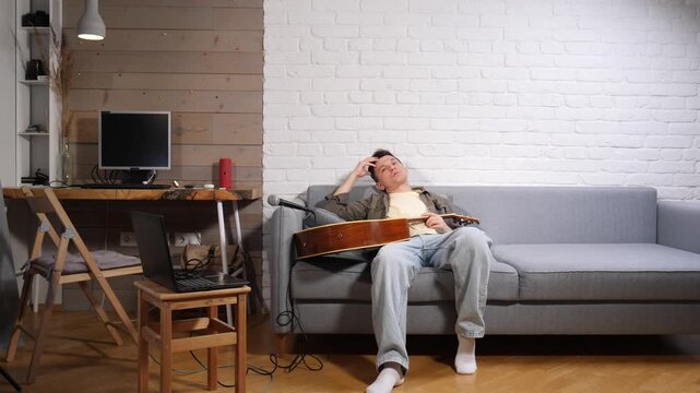 Exhausted musician sitting on a sofa with his acoustic guitar, experiencing creative burnout after spending time in his home studio, needing a rest and contemplating his work