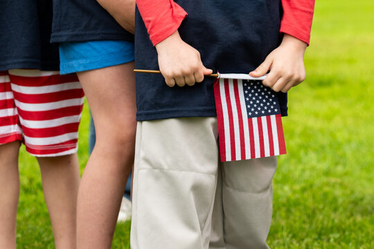 A child's hands grip a small American flag, baseball pants visible in the frame, combining patriotism and America's pastime in one image.