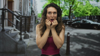 Fototapeta premium Young hispanic woman holds fingers near chin with open mouth on bustling city street; surprise.