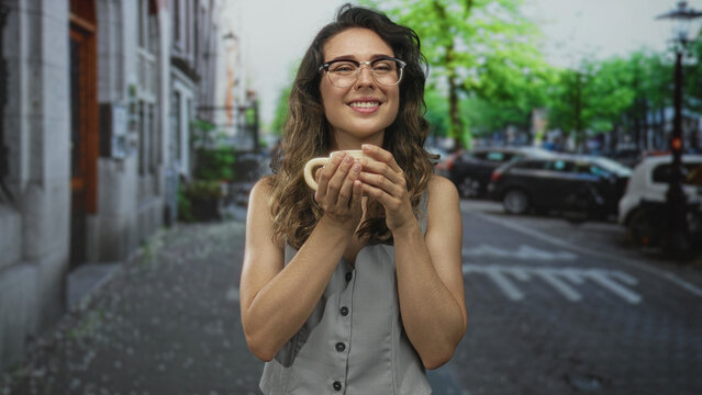 Young hispanic woman wearing glasses holds a ceramic coffee mug while smiling on a city street; calm reflection.
