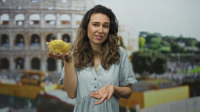 Young hispanic woman holds bowl of tortilla chips with a shrug gesture at the coliseum building; indifference.